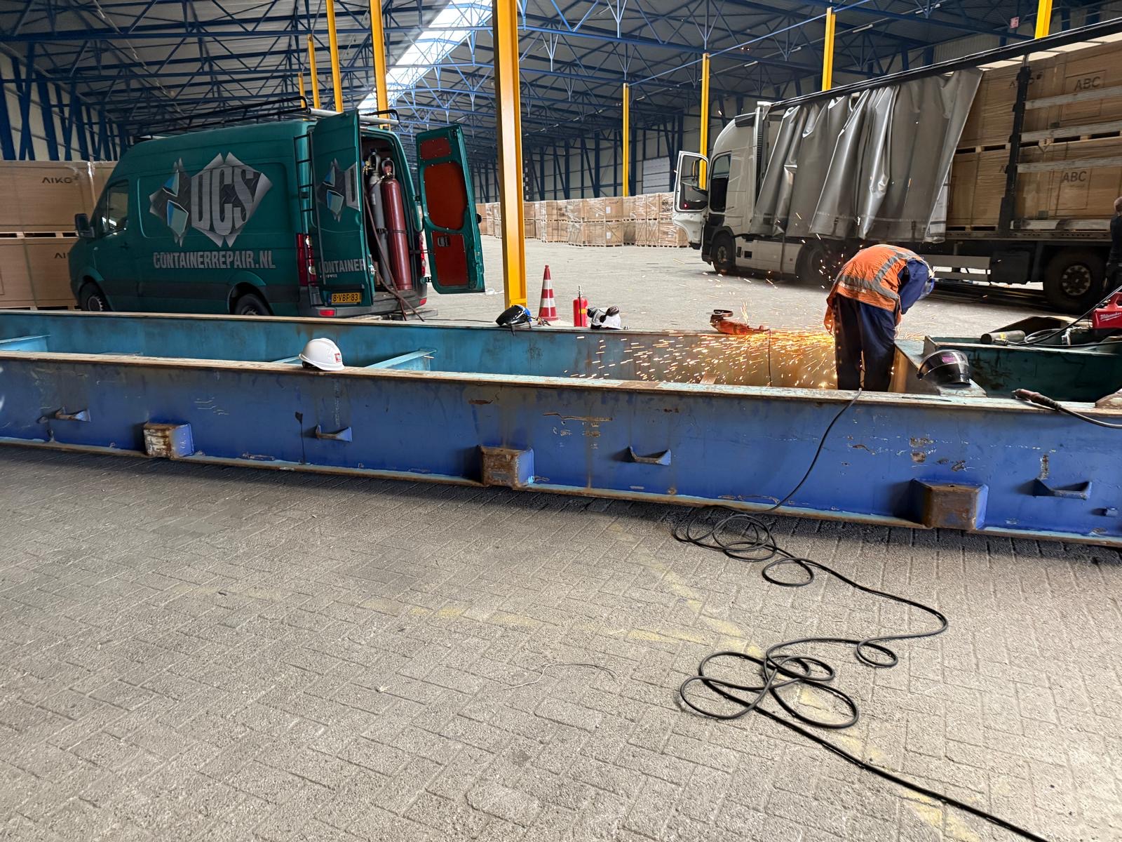 DCS servicebus met containerrepair.nl-opdruk naast een blauw flatrack tijdens laswerkzaamheden in de werkplaats
